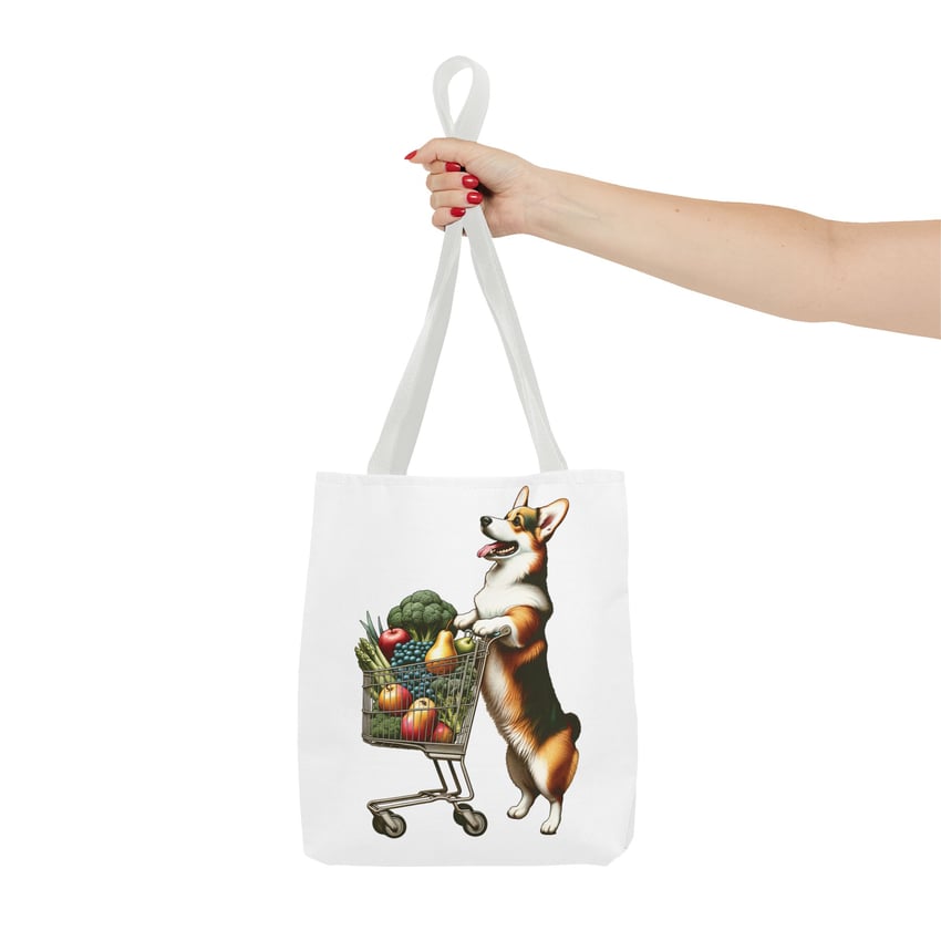 White tote bag with playful corgi illustration and fresh produce, held by hand with red nails.